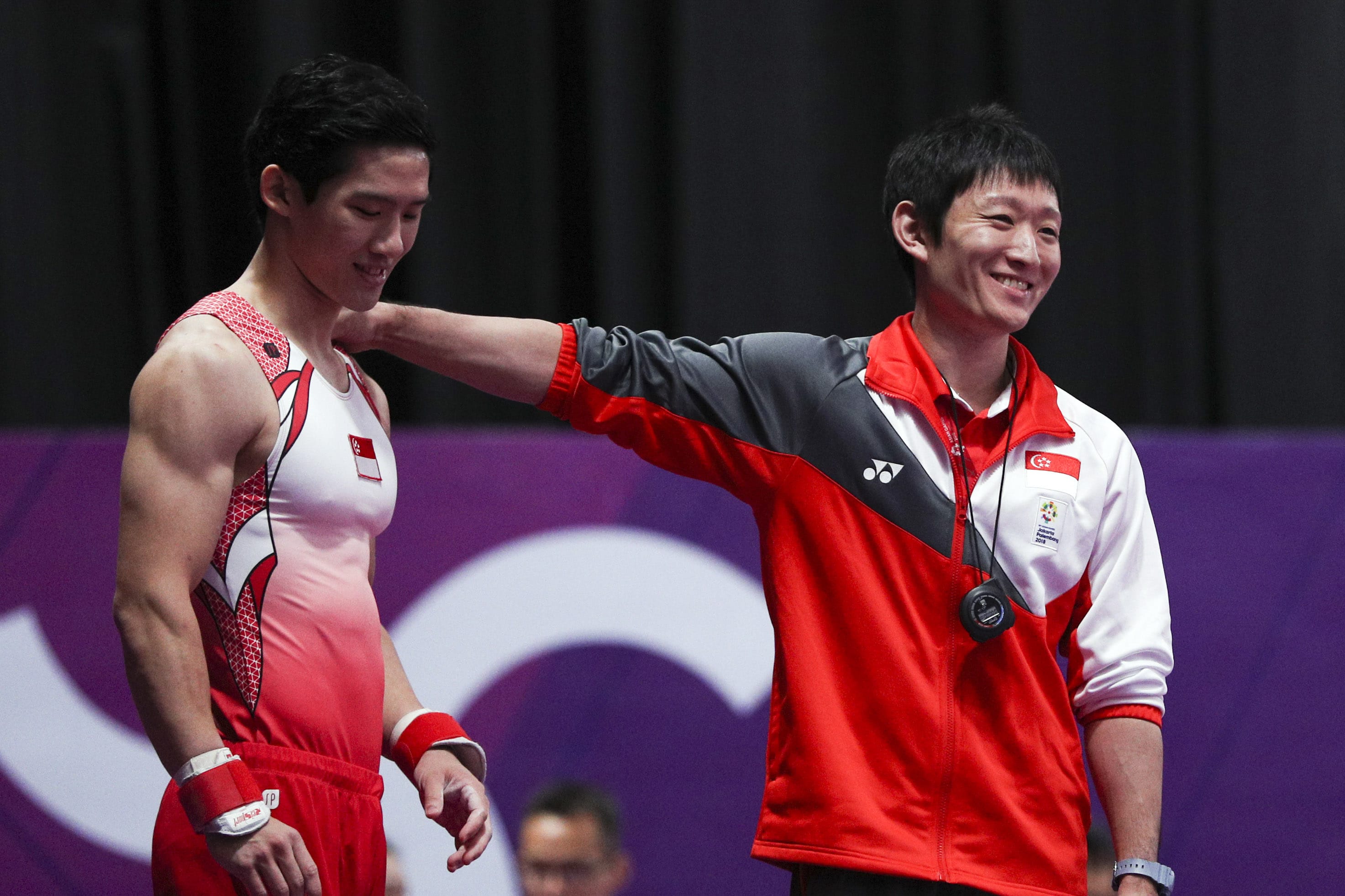 Two smiling male athletes in red-and-white uniforms pose on a purple podium backdrop; one extends his arm across the other’s shoulders in a friendly, celebratory moment.